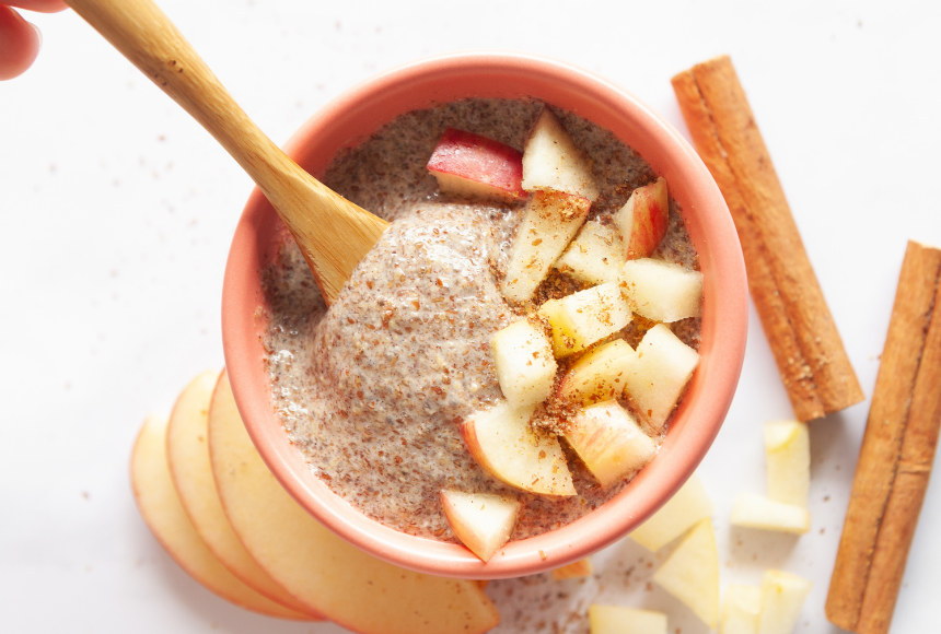 apple cinnamon superseed pudding displayed in an orange bowl with a small wooden spoon, beside the bowl are cinnamon sticks, apple slices and sprinkles of cinnamon