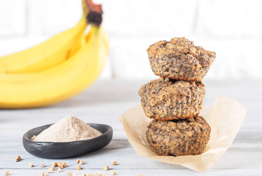 banana chia muffins stacked on top of each other in a paper muffin wrapper, displayed behind the muffins are a couple bright yellow bananas and some ground cinnamon in a small black bowl with oats scattered around