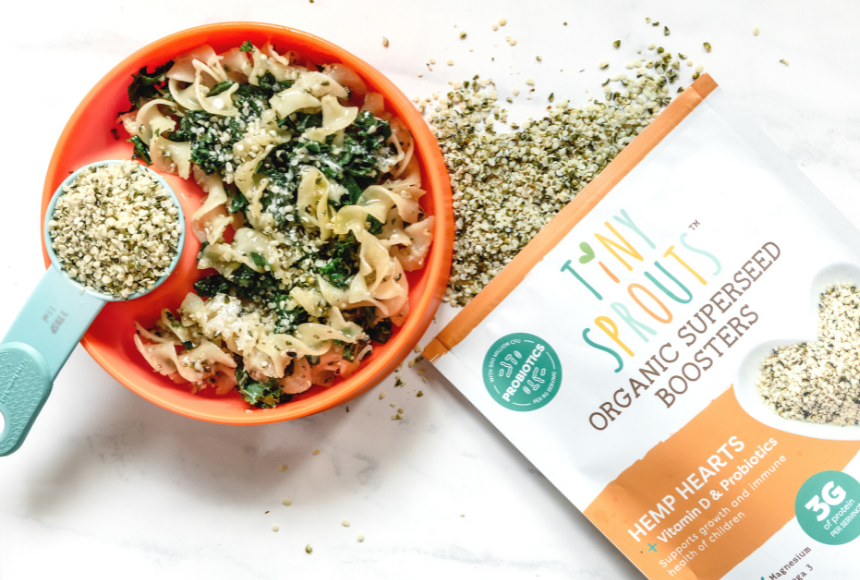 Buttered noodles with kale and hemp hearts in a red bowl on a marble countertop with a bag of Tiny Sprouts Organic Hemp Hearts beside it