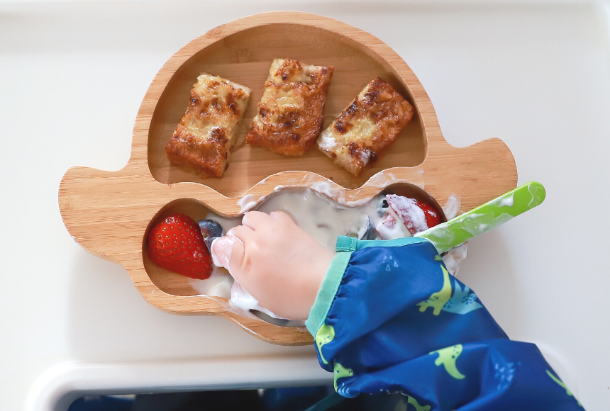 Flaxseed french toast bites with a wooden plate with strawberries and yogurt. Toddler hand is grabbing the fruit and yogurt.