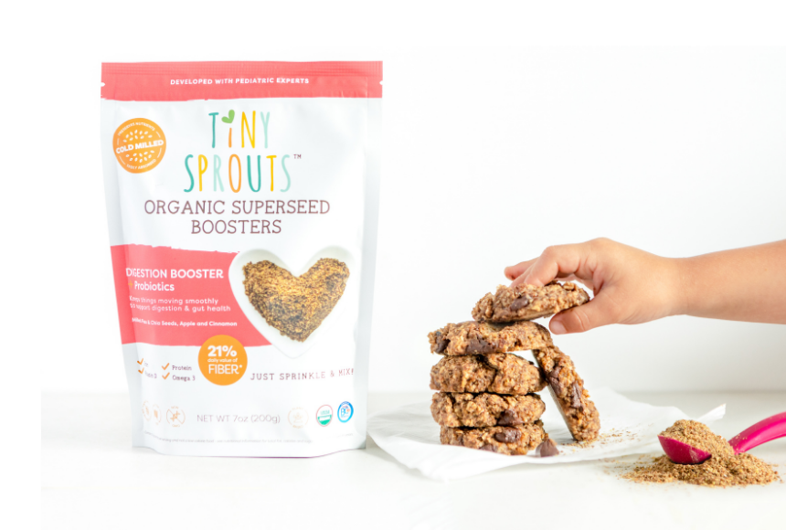 Stack of Apple Drop Digestive cookies on a white napkin with a toddlers hand reaching for the top cookie in the stack. Stack is beside a bag of Tiny Sprouts Organic Digestion Booster