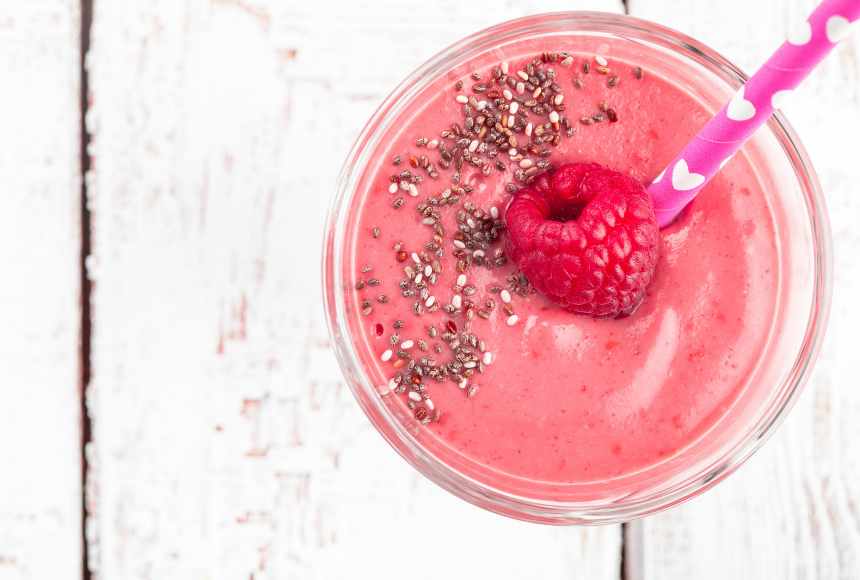 Top view of raspberry chia smoothie in a clear glass with pink and white heart straw on a white wooden table
