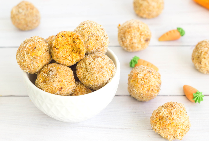 carrot cake energy balls placed in a small white bowl with one half eaten energy bite on top, displayed around the bowl are scattered carrot energy balls and mini carrots