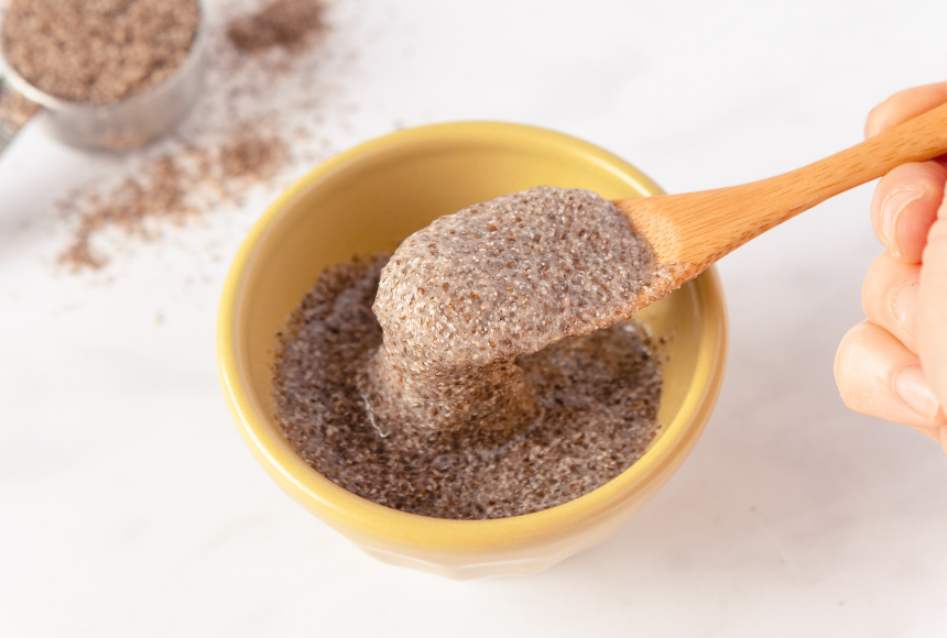chia egg displayed in a small yellow bowl with an orange spoon, displayed beside the bowl is a small measuring cup of chia seeds with chia seeds scattered around the measuring cup