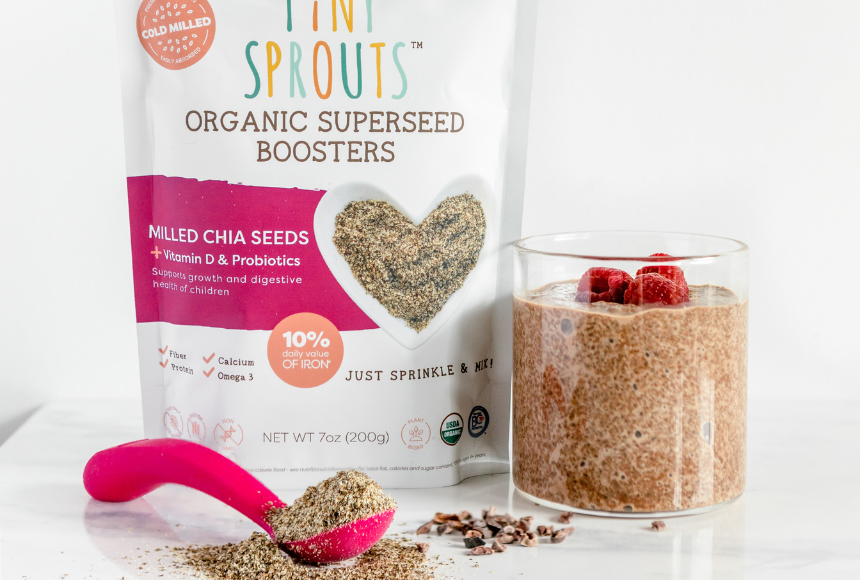 Glass of chocolate chia pudding with raspberries on top beside a bag of Tiny Sprouts Organic Milled Chia seeds and a pink spoon of milled chia seeds in front 