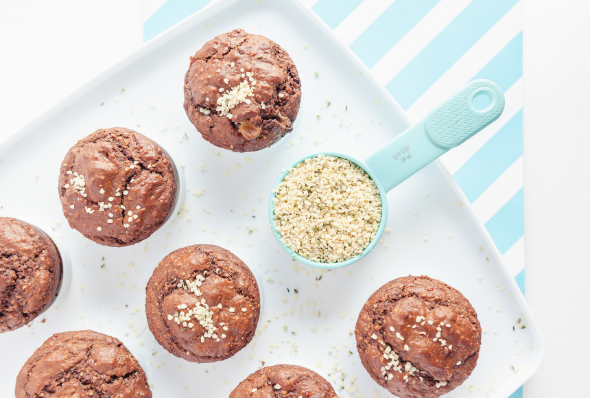 chocolate protein muffins sprinkled with hemp hearts on a muffin tray with a light blue measuring cup of hemp hearts 