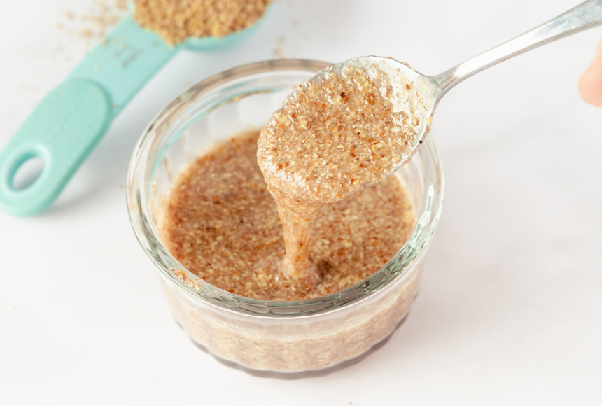 flax egg displayed in a small glass bowl with a silver spoon, beside the bowl is a small blue measuring spoon full of flax seeds with some flax seeds scattered around