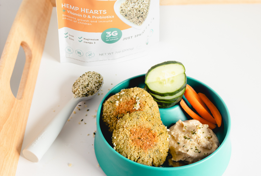 Hemp heart falafels in a teal bowl with hummus, cucumber and carrots with a spoon of hemp hearts beside it