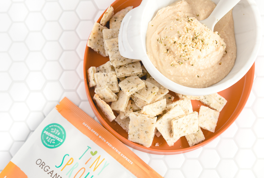 Hemp heart hummus in white bowl with hemp seeds sprinkled on top. Bowl of hummus is on an orange plate with baked pita bread