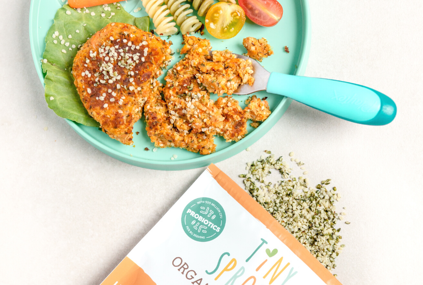 Hemp veggie burger made on a teal plate topped with hemp seeds and beside tomatoes and pasta. Plate is beside am open bag of Tiny Sprouts Organic Hemp Hearts