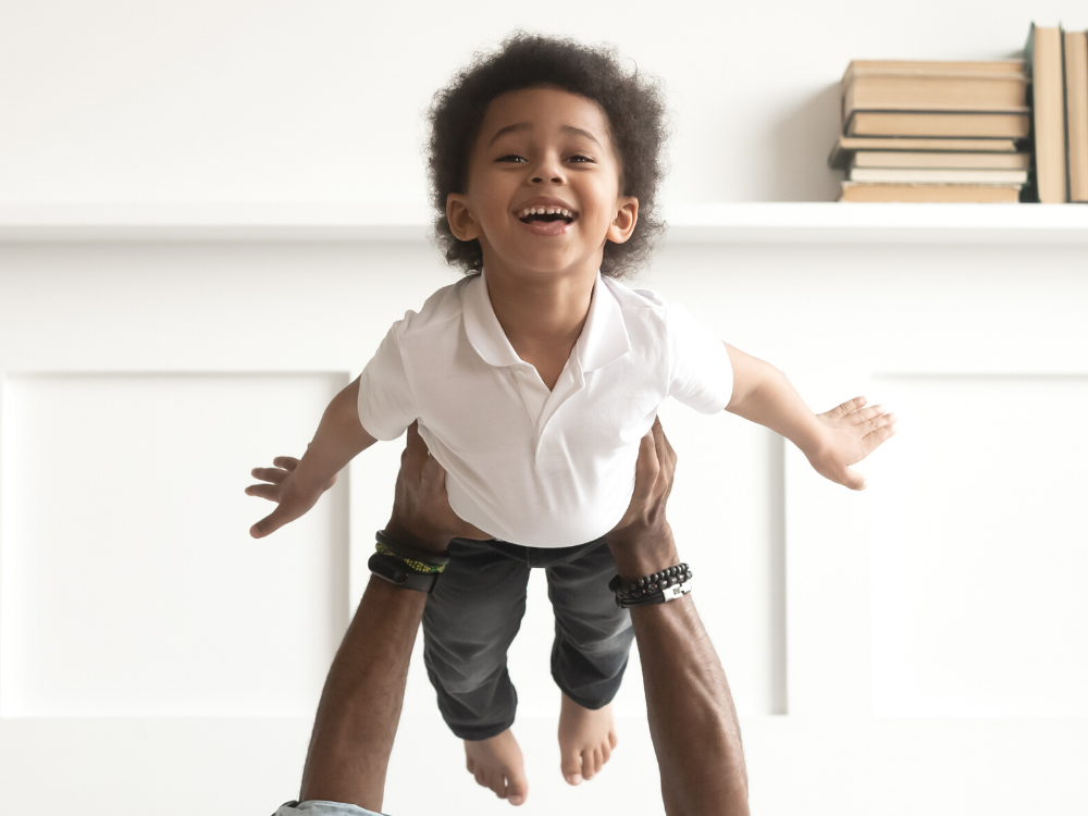A happy and cute African American toddler being lifted in the air, posing like he is flying to resemble a 'Superkid'.